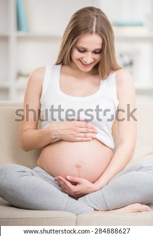 Beautiful pregnant woman keeping her hands on belly while sitting at sofa.