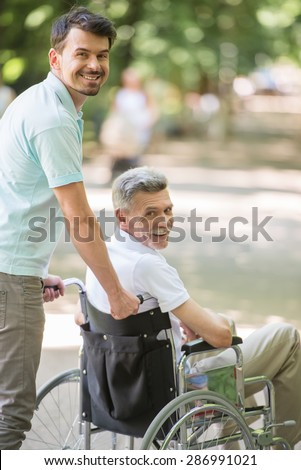 Adult son walking with disabled father in wheelchair at park.