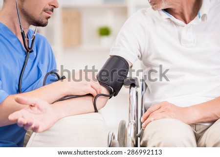 Male doctor measuring blood pressure to older patient sitting at wheelchair.