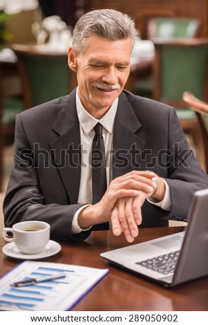 Elegant mature businessman sitting at the table in cafe and looking at watches.