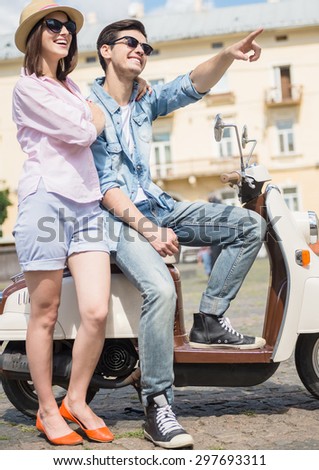 Beautiful young couple dressed casual sitting on scooter together, man pointing away.