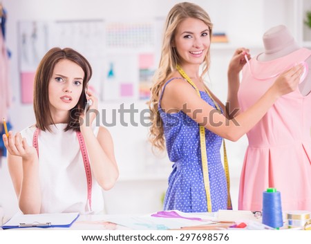 Blond hair woman working at pink dress on mannequin and brown hair woman speaking by phone in studio.