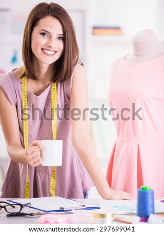 Beautiful young female fashion designer holding cup of tea and smiling at camera, mannequin on background.