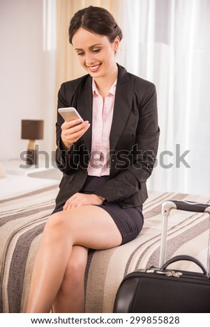 Cheerful business woman looking on phone while sitting on bed at the hotel room.