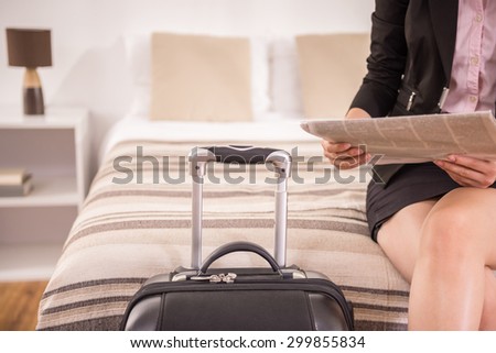 Business woman holding newspaper while sitting on bed near suitcase at the hotel room. Close-up.