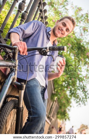 Young beautiful man standing with bicycle at the city street near the fence with cup of coffee.