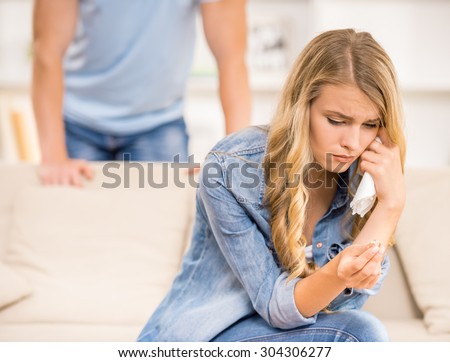 Crying woman sitting on sofa with wedding ring while husband standing behind her.