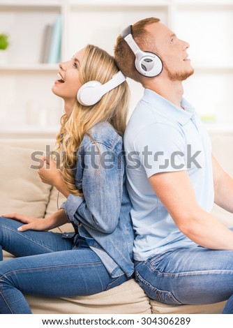 Beautiful couple listen to music with headphones while sitting back to back at home on the sofa.