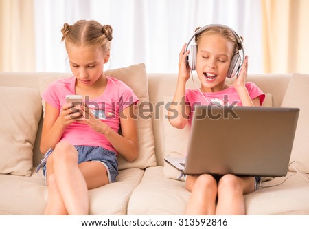 Young twins sisters are using laptop, headphone and smartphone sitting on the couch.