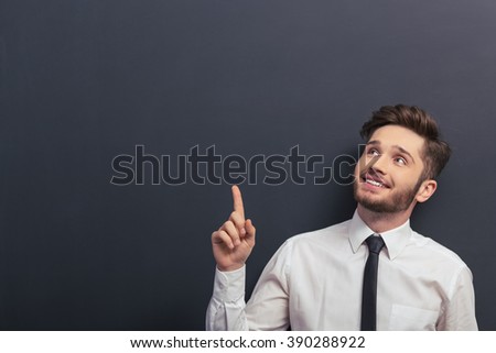 Handsome young student in white classic shirt is smiling, looking and pointing up, standing against blackboard