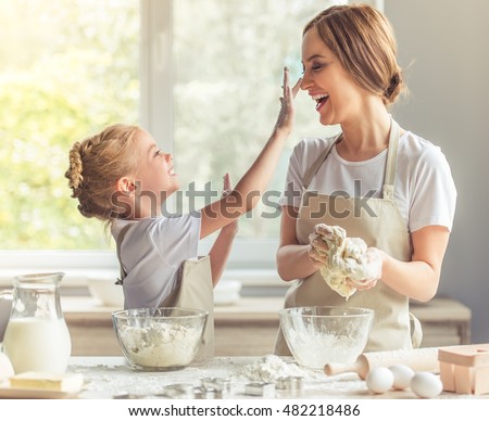 Cute little girl and her beautiful mom in aprons are playing and laughing while kneading the dough in the kitchen