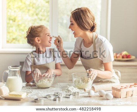 Cute little girl and her beautiful mom in aprons are playing and laughing while kneading the dough in the kitchen