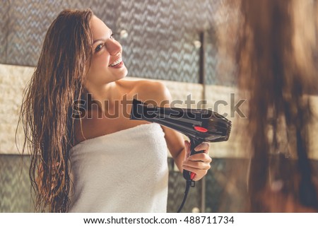 Beautiful young woman in bath towel is using a hair dryer and smiling while looking into the mirror in bathroom
