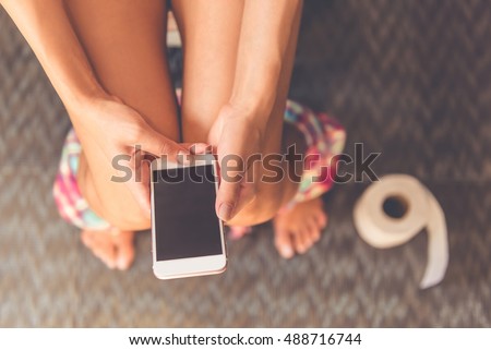 Cropped image of beautiful young woman using a smartphone while sitting on toilet, close-up