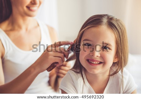 Charming little girl is looking at camera and smiling while her beautiful young mother is combing daughter\'s hair