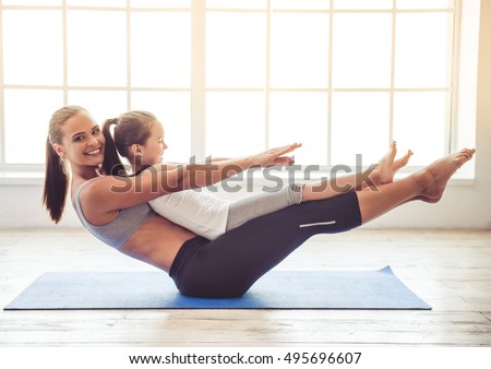 Beautiful young woman and charming little girl are smiling while doing yoga together in fitness hall