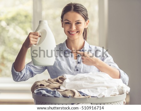 Beautiful young woman is holding a fabric softener over the basin with laundry, looking at camera and smiling