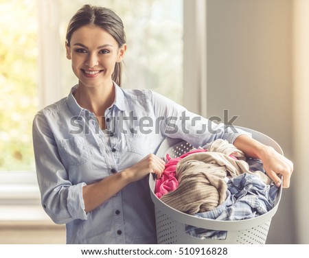 Beautiful young woman is holding a basin with laundry, looking at camera and smiling while standing at home