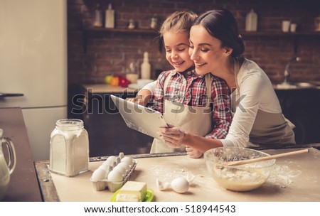 Beautiful young mom and her cute little daughter are using a digital tablet and smiling while baking in kitchen at home