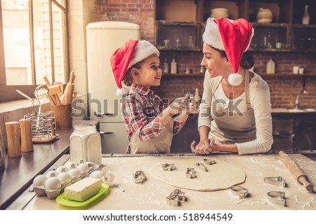 Beautiful young mom and her cute little daughter in Santa hats are using cookie cutters and smiling while baking in kitchen at home
