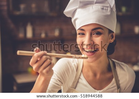 Beautiful young woman in chef hat is holding wooden spoon, looking at camera and smiling