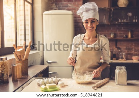 Beautiful young woman in chef hat is mixing batter, looking at camera and smiling while baking in kitchen at home