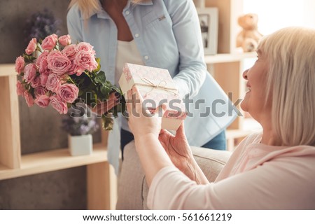 Beautiful adult woman is giving flowers and a gift box to her mature mother and smiling while sitting on couch at home