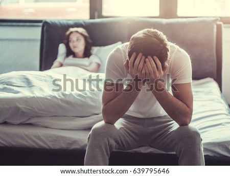We have a problem. Upset young man sitting on the edge of the bed and holding his head against the background of a girlfriend, lying in bed.