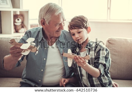 Grandpa and grandson are playing with toy planes and smiling while sitting on couch at home