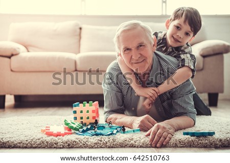 Grandpa and grandson are playing with toys, hugging, looking at camera and smiling while resting together at home
