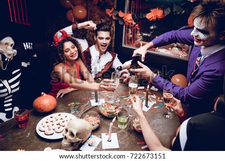 Halloween party. The guy in the Joker costume spills his champagne glasses in glasses. They are sitting at a table in a nightclub for Halloween. On the table are sweets