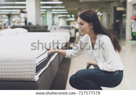A woman in a white shirt and jeans in a mattress store. She examines the mattress she wants to buy. She squats and looks at the mattress