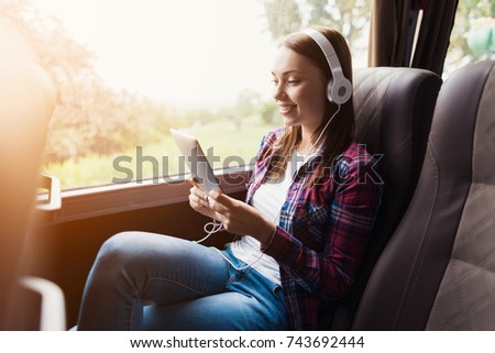 The woman on the passenger seat of the bus listens to music and looks at the tablet. She looks at the device\'s screen and smiles. Outside the window is a beautiful green landscape.