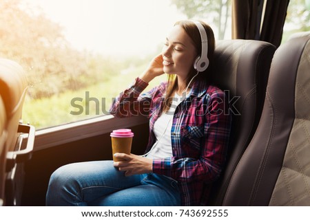 The woman on the passenger seat of the bus listens to music and drinks coffee. She looks out the window and smiles. Outside the window is a beautiful green landscape.