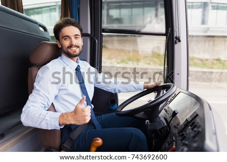 The driver of the tourist bus is smiling and looking at the camera. He is posing behind the wheel of a bus in the driver\'s seat. The man runs a new black tourist bus.