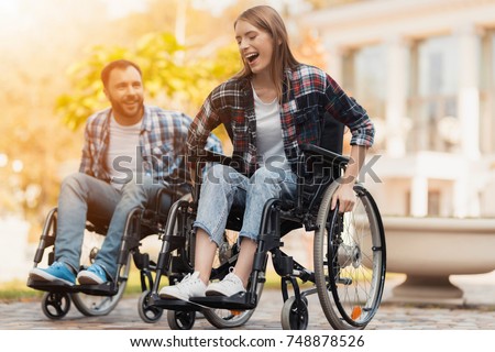 A man and a woman on wheelchairs ride around the park. They arranged a race in wheelchairs. On a background of a beautiful green park.