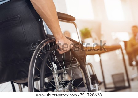 A disabled man is sitting in a wheelchair. He holds his hands on the wheel. Nearby are his colleagues