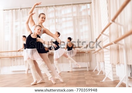 The trainer of the ballet school helps young ballerinas perform different choreographic exercises. They rehearse in the ballet class. The teacher communicates with the children.