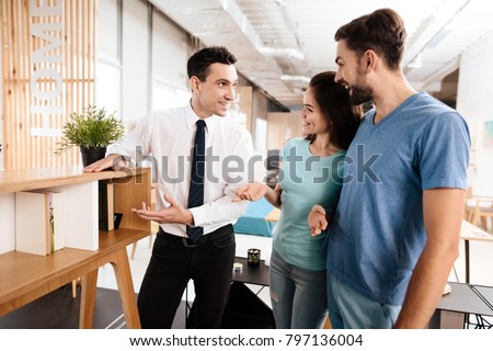 The furniture store manager demonstrates furniture to the buyers. A guy and a girl are listening with a good mood to the seller.