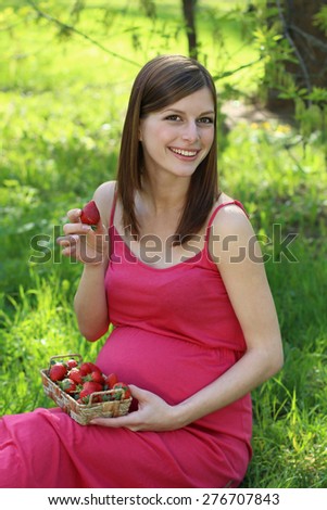 Beautiful young pregnant woman sitting on green grass with juicy strawberry in hands