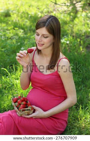 Beautiful young pregnant woman sitting on green grass with juicy strawberry in hands