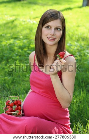 Pregnant woman sitting on green grass with juicy strawberry in hands