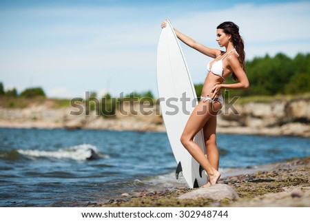 Sexy brunette girl in white swimsuit posing with a surfing board on a beach