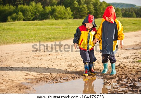 Two boys walking through a mud puddle in her rain coat and boots.