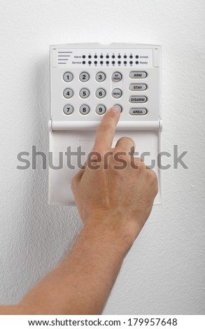 A regular security systems keypad with buttons being pressed by a mans hand on a white textured wall