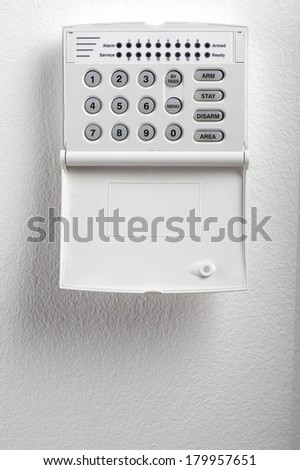 A regular security systems keypad with buttons and a plastic flap mounted on a white textured wall