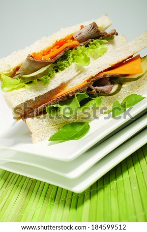 A closeup of a beef sandwich made with white bread, gherkins, lettuce and cheese on white plates and a green bamboo mat