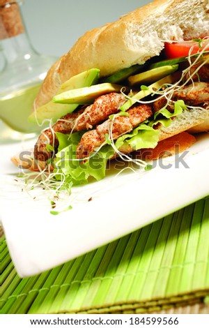 A chicken sandwich made with chicken strips, avocado lettuce and sprouts on a white plate  on a green bamboo mat