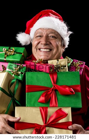 Jovial male senior with broad grin is peering across a load of presents that he is carrying against his chest. Gifts wrapped in plain red, green, magenta and gold. Red Father Christmas cap and coat.