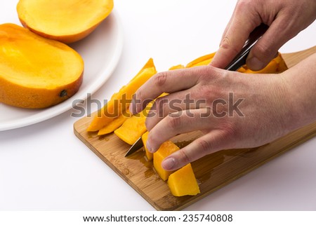 Two hands cutting mango chips into dice. The fingers of a left hand are fixing the fruit flesh on a wooden board, while the right is guiding a knife. Two mango slices on a plate. White background.